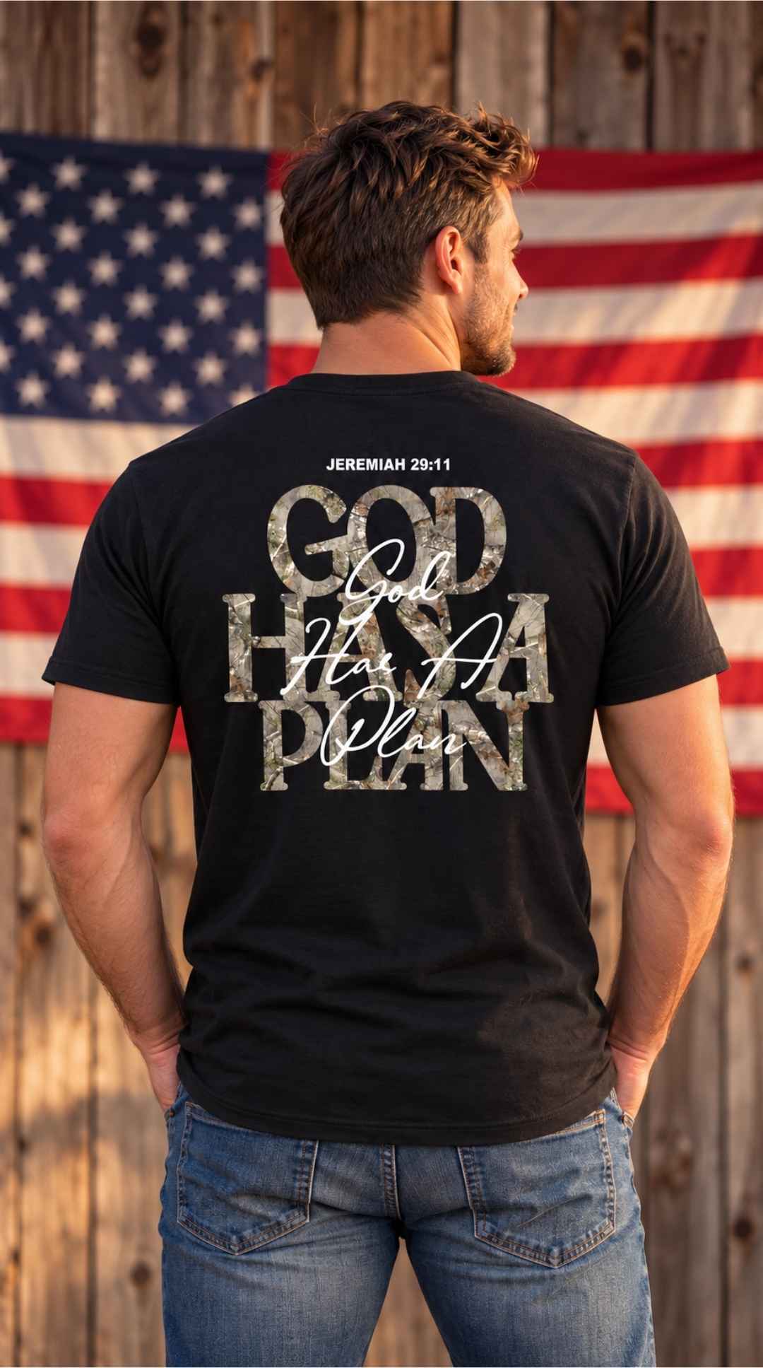 A man viewed from behind wearing a black graphic t-shirt with the text 'GOD HAS A PLAN' in distressed white and gray lettering, standing in front of a rustic wooden fence and an American flag.